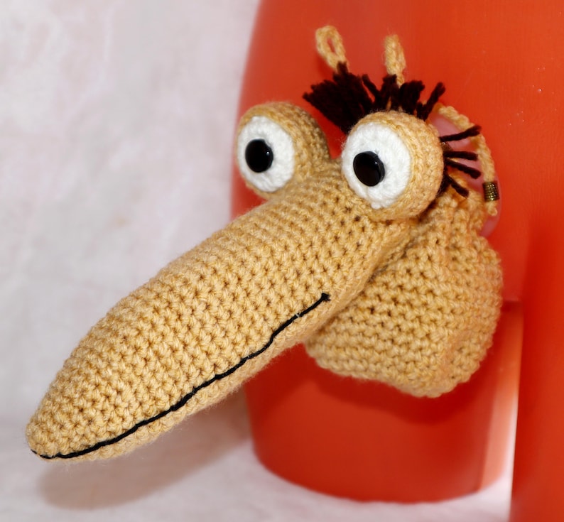May include: A whimsical, handmade crochet creature with a long, tan snout, large white-rimmed eyes with black pupils, and a small tuft of dark brown hair. The creature is attached to an orange surface.