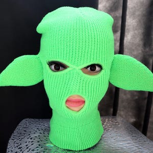 May include: A bright neon green knit balaclava featuring two large, pointed, ear-like protrusions extending from the sides. The head covering has two eye openings and one mouth opening, revealing a face with dark eyelashes and pink lips.