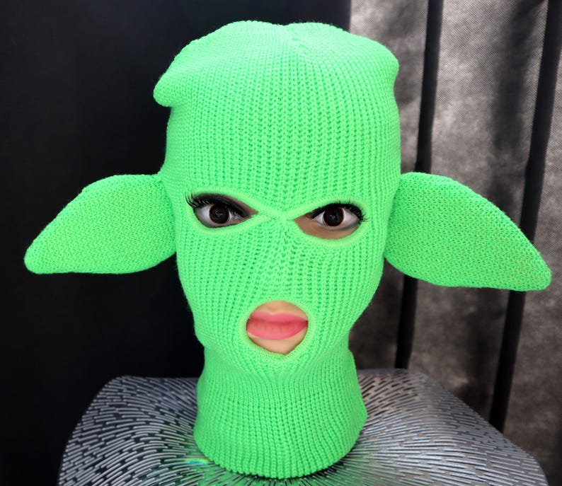 May include: A bright neon green knit balaclava featuring two large, pointed, ear-like protrusions extending from the sides. The head covering has two eye openings and one mouth opening, revealing a face with dark eyelashes and pink lips.