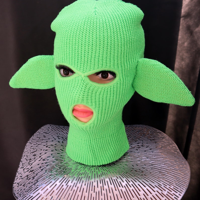 May include: A bright neon green knit balaclava featuring two large, pointed, ear-like extensions on the sides. The head covering has three distinct openings for the eyes and mouth, revealing a face underneath with visible eyelashes and pink lips.