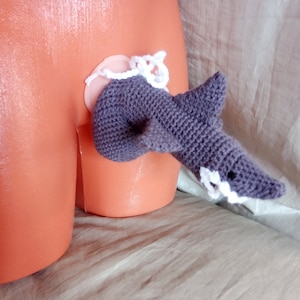 May include: A gray crocheted shark toy with white crocheted details. The toy is attached to a pink and white crocheted ring.