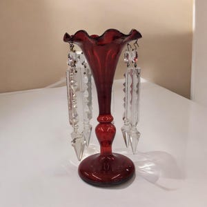 May include: A ruby red glass vase with a flared, ruffled top and a round base. Clear crystal prisms hang from the rim. The vase has a slender neck and a bulbous midsection, adding a touch of elegance.