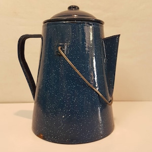May include: A dark blue enamel coffee pot with a white speckled pattern, featuring a matching lid, a side handle, a metal bail handle, and a spout. The surface shows signs of wear with minor paint loss near the base and on the metal handle.