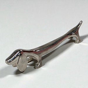 May include: A silver-colored dachshund dog figurine with a long body, short legs, and a curved tail. The dog has a detailed head with a prominent ear and a smooth, reflective surface. The figurine is likely made of metal.