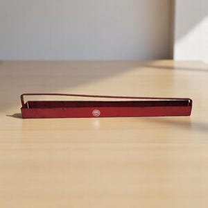 May include: A long, rectangular red metal tray with a handle. The tray has a slightly distressed finish and a small circular logo on the front. The handle is attached to the top of the tray. The tray is sitting on a light-colored wooden surface.