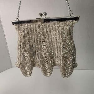 May include: Silver-toned vintage handbag with a chain strap. The bag features a decorative, draped design. The clasp is a silver metal frame with two round ball closures. The bag's body is made of small, linked metal pieces.