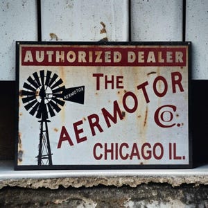 May include: Vintage metal sign with the text "AUTHORIZED DEALER THE AERMOTOR @ C. CHICAGO IL." The sign features a black windmill graphic and is framed in black. The sign has a distressed, aged appearance.