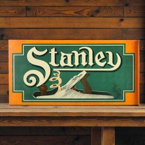 May include: A rectangular sign featuring the word "Stanley" in a vintage font, with a green background and an orange border. The sign also depicts a hand plane. The sign is displayed on a wooden shelf.