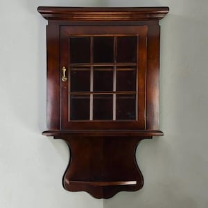 May include: A dark brown wooden wall-mounted cabinet featuring a single door with a nine-pane glass window and a small brass handle. The unit includes a decorative crown molding at the top and an open shelf at the bottom with a curved base design.