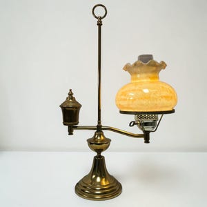 May include: An antique brass table lamp with a decorative design. The lamp features a round base, a central vertical support, and a curved arm holding a yellow glass shade. A smaller lamp is on the left side. The lamp has a ring at the top.