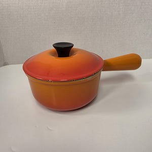 May include: Orange enameled cast iron saucepan with a lid and wooden handle. The lid has a black knob. The saucepan is designed for cooking and has a classic, vintage look. The handle is approximately 6 inches long.