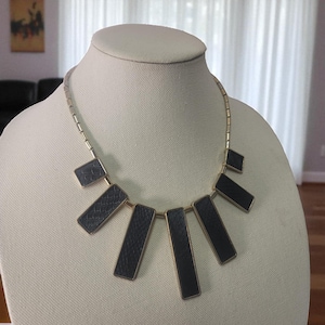 May include: A gold-toned necklace with a series of dark gray rectangular and diamond-shaped pendants. The pendants are arranged in a fan-like design. The necklace is displayed on a mannequin.