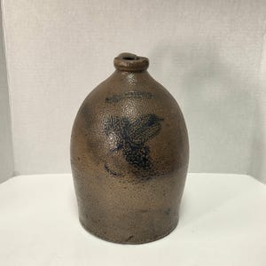 May include: Brown stoneware jug with a rounded body and a narrow neck. The jug features a dark blue floral design on the front. The surface has a textured appearance, and the jug appears to be an antique or vintage piece.