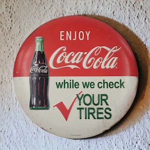 May include: A vintage Coca-Cola sign with a red and white circular design. The sign features a Coca-Cola bottle graphic and the text "ENJOY Coca-Cola while we check YOUR TIRES". The sign has a distressed, aged appearance.