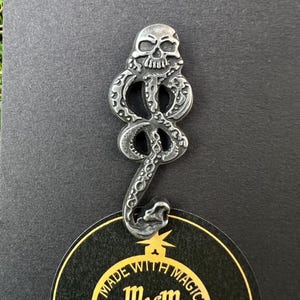 Pode incluir: Um alfinete prateado com um design de caveira sobre uma cobra. O alfinete está preso a um cartão preto com um rótulo dourado que diz "MADE WITH MAGICS" e "MWM".