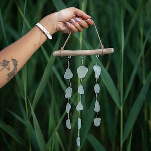 Puede incluir: Una mano sostiene un carillón de madera flotante con una cadena de vidrio de mar colgando de él. El carillón está hecho de madera flotante natural y tiene un aspecto rústico. El vidrio de mar es transparente y tiene un acabado liso y pulido.