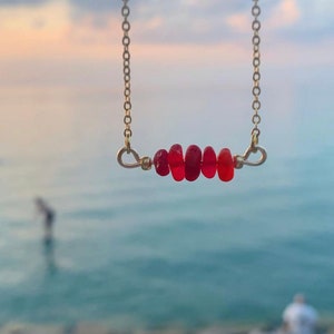 May include: A gold chain necklace with a row of five red glass beads. The necklace is photographed against a blurred background of a beach and ocean.