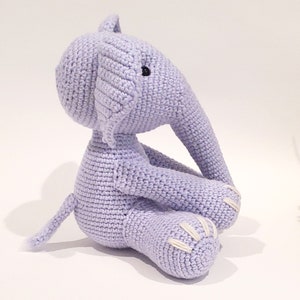 May include: A crocheted lavender elephant plush toy sitting with its legs crossed. The elephant has white stitching on its feet and a black button eye.