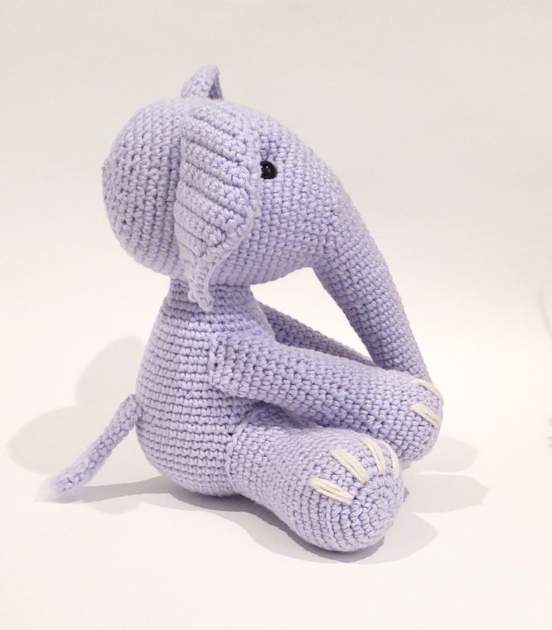 May include: A crocheted lavender elephant plush toy sitting with its legs crossed. The elephant has white stitching on its feet and a black button eye.