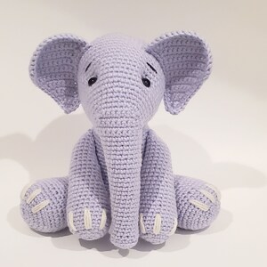 May include: A crocheted lavender elephant plush toy with white accents on the feet and ears. The elephant is sitting with its legs crossed and has black eyes.
