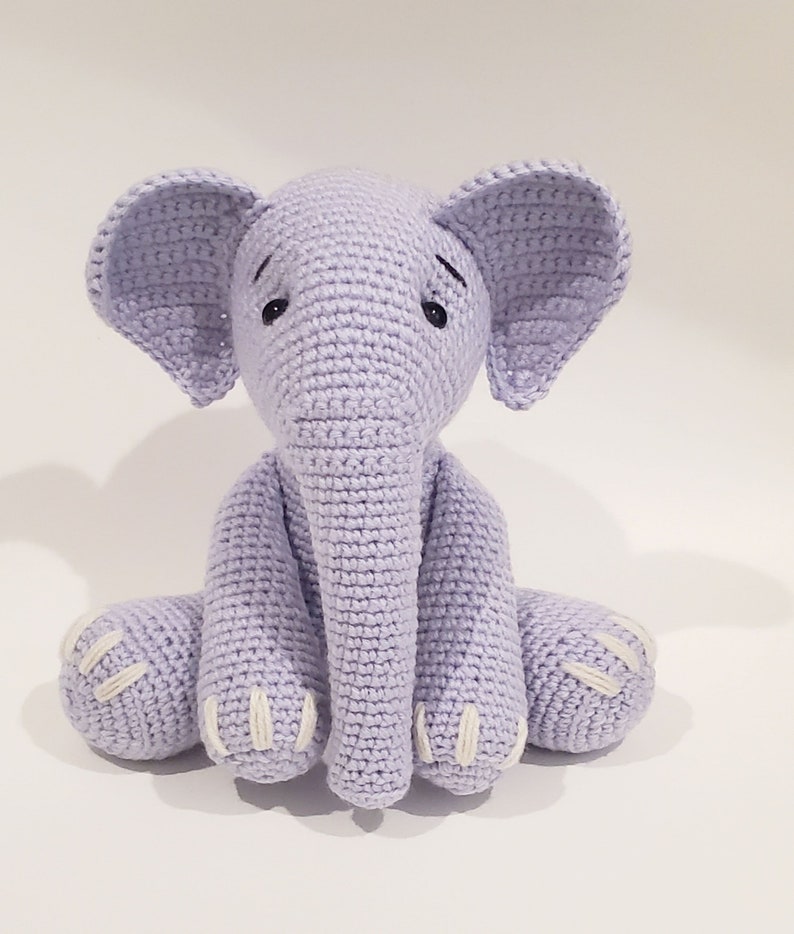 May include: A crocheted lavender elephant plush toy with white accents on the feet and ears. The elephant is sitting with its legs crossed and has black eyes.