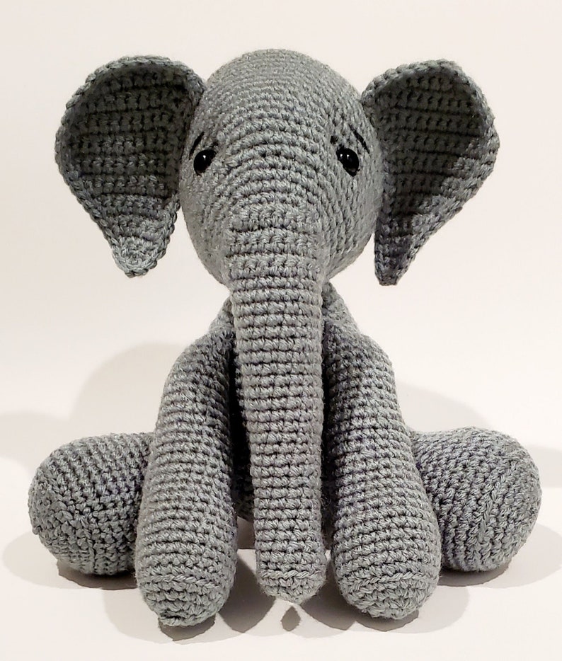 May include: A gray crocheted elephant plush toy with large ears and a long trunk. The elephant is sitting with its legs crossed.