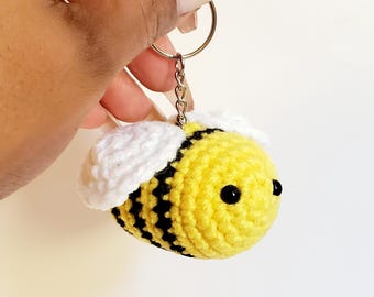 May include: A handmade crochet bee keychain. The bee is yellow with black stripes and white wings. It has black eyes and is attached to a silver keyring. A cute and whimsical accessory.