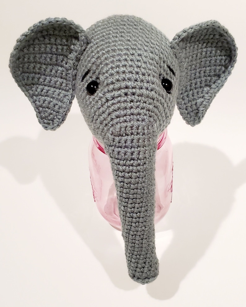 May include: A gray crocheted elephant head with black eyes, sitting on top of a pink glass jar.