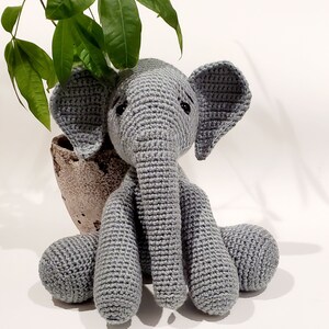 May include: A gray crocheted elephant toy sitting on a white surface. The elephant has a long trunk and large ears.