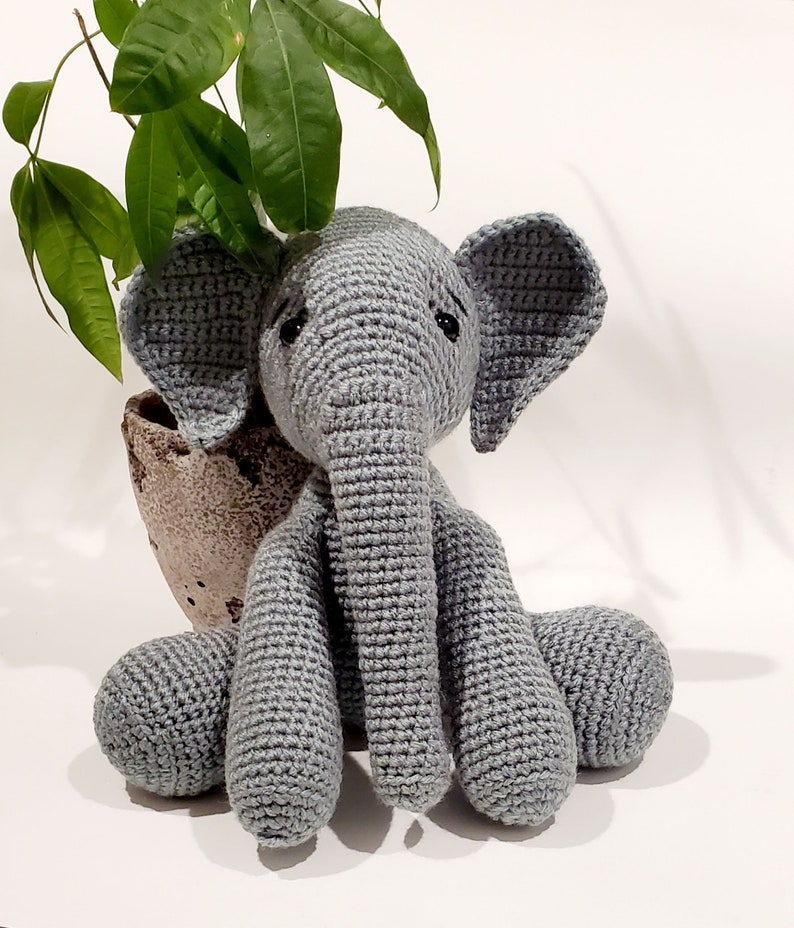 May include: A gray crocheted elephant toy sitting on a white surface. The elephant has a long trunk and large ears.
