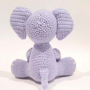 May include: A crocheted lavender elephant toy sitting on a white surface. The elephant has a long trunk and large ears.