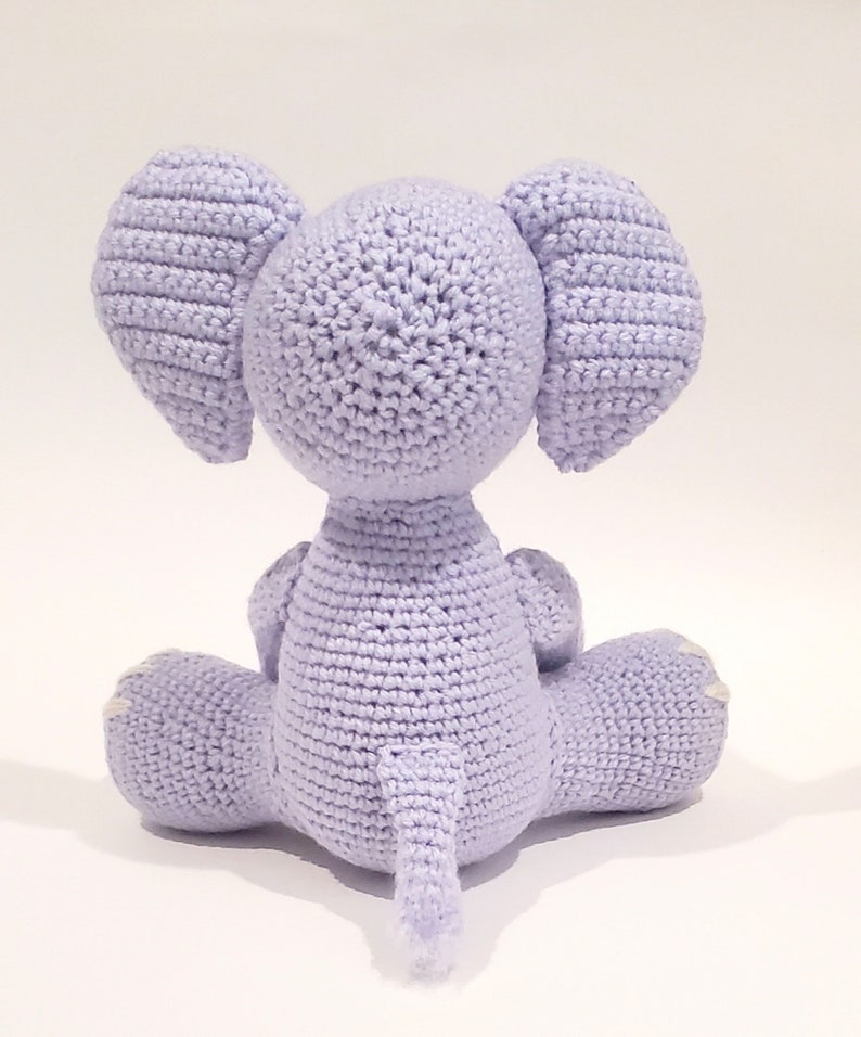May include: A crocheted lavender elephant toy sitting on a white surface. The elephant has a long trunk and large ears.