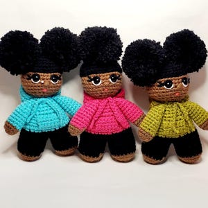 May include: Three handmade crochet dolls with brown skin, black yarn hair styled in two large pom-pom buns, and embroidered facial features. Each doll wears black pants and a hooded sweatshirt in bright turquoise, hot pink, or olive green.