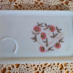 May include: A rectangular, white serving tray with a scalloped edge and handles. The tray features a floral design with pink flowers and brown leaves. A curved indentation is on the left side of the tray.