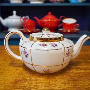 May include: A white porcelain teapot with a gold rim and accents, featuring a floral pattern. The teapot has a curved handle and spout. Other teacups and teapots are visible in the background, suggesting a tea set.