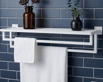 May include: A white metal wall-mounted bathroom shelf featuring a top rack and a lower towel bar. The shelf holds a brown glass bottle with dried lotus pods and a small black vase with green foliage. A white textured towel hangs from the bar.