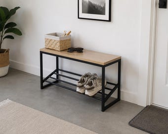 May include: A rectangular hallway bench featuring a light wood grain top surface and a black metal frame. The bench includes a lower slanted shelf with three horizontal black metal bars for shoe storage, currently holding a pair of beige trainers.