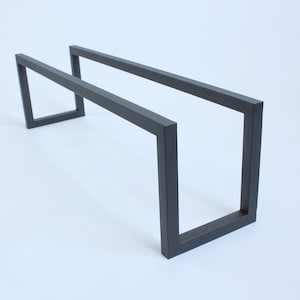 May include: A matte black metal frame featuring a rectangular, open-ended design with two parallel horizontal bars connected by two vertical U-shaped supports at each end, creating a hollow, elongated structure with clean, sharp edges.