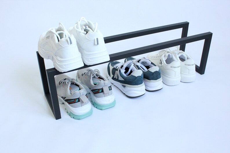 May include: A black metal shoe rack with a two-tier design, holding four pairs of athletic trainers. The top tier features two white trainers, while the bottom tier holds two pairs of grey and teal athletic trainers and white trainers.