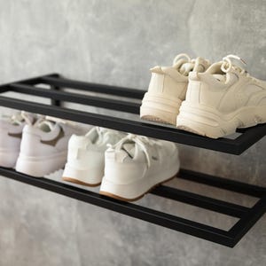 Może przedstawiać: Dwupoziomowy, ścienny stojak na buty wykonany z czarnego metalu o prostokątnej ramie. Na g&oacute;rnej p&oacute;łce znajduje się para kremowych sneakers&oacute;w, natomiast na dolnej p&oacute;łce umieszczono dwie pary białych sneakers&oacute;w z grubymi podeszwami i sznurowadłami.