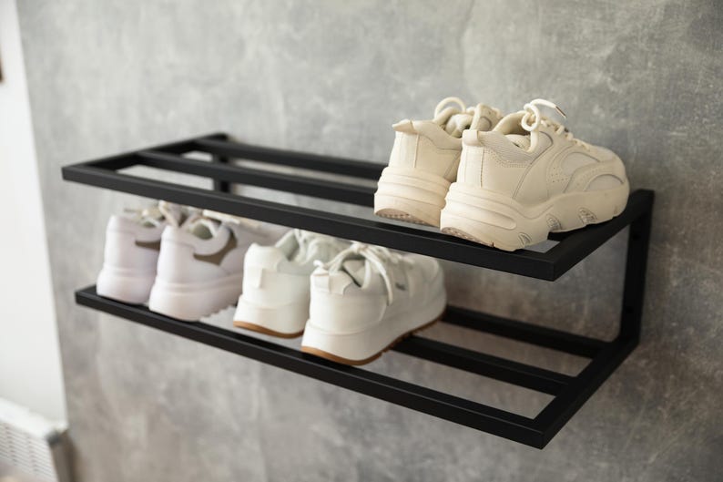 Może przedstawiać: Dwupoziomowy, ścienny stojak na buty wykonany z czarnego metalu o prostokątnej ramie. Na g&oacute;rnej p&oacute;łce znajduje się para kremowych sneakers&oacute;w, natomiast na dolnej p&oacute;łce umieszczono dwie pary białych sneakers&oacute;w z grubymi podeszwami i sznurowadłami.