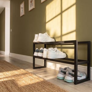 May include: A black metal shoe rack with two horizontal tiers. The top shelf holds a pair of white trainers, while the middle shelf holds two pairs of white trainers. The bottom shelf holds one pair of white and light blue trainers.