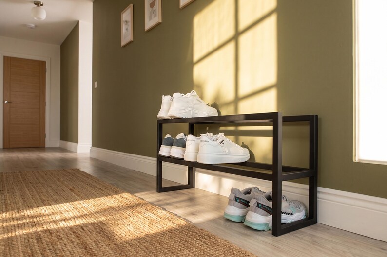 May include: A black metal shoe rack with two horizontal tiers. The top shelf holds a pair of white trainers, while the middle shelf holds two pairs of white trainers. The bottom shelf holds one pair of white and light blue trainers.