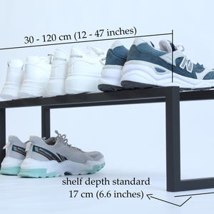 May include: A black metal shoe rack with a rectangular frame. The rack measures 30-120 cm (12-47 inches) in width, 20 cm (8 inches) in height, and has a standard shelf depth of 17 cm (6.6 inches). Several pairs of athletic trainers are displayed.