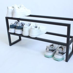 May include: A black metal shoe rack with two tiers, featuring a rectangular frame design. The top tier holds a pair of white trainers, the middle tier holds two pairs of trainers, and the bottom tier holds a pair of grey and teal trainers.