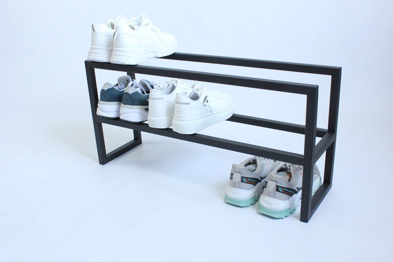 May include: A black metal shoe rack with two tiers, featuring a rectangular frame design. The top tier holds a pair of white trainers, the middle tier holds two pairs of trainers, and the bottom tier holds a pair of grey and teal trainers.
