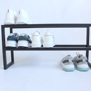 Op de afbeelding: Een zwart metalen schoenenrek met twee horizontale niveaus. Het bovenste niveau bevat een paar witte sneakers. Het onderste niveau bevat een paar blauw-witte sneakers, een paar witte sneakers en een paar grijze sneakers met lichtblauwe zolen.