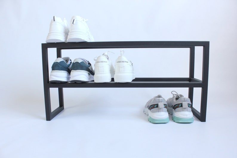 Op de afbeelding: Een zwart metalen schoenenrek met twee horizontale niveaus. Het bovenste niveau bevat een paar witte sneakers. Het onderste niveau bevat een paar blauw-witte sneakers, een paar witte sneakers en een paar grijze sneakers met lichtblauwe zolen.