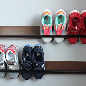 May include: Two black metal shelves with a wood grain finish, mounted on a white wall. The shelves are holding six pairs of sneakers in various colors, including red, blue, gray, and white.