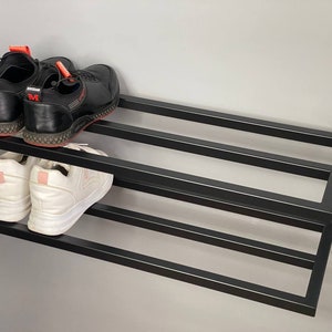 Może przedstawiać: Ścienny stojak na buty z czarnego metalu z dwoma poziomami. Na g&oacute;rnym poziomie znajduje się para czarnych sneakers&oacute;w z czerwonymi akcentami, a na dolnym para białych sneakers&oacute;w z jasnor&oacute;żowymi zapiętkami, o konstrukcji z poziomych listew.